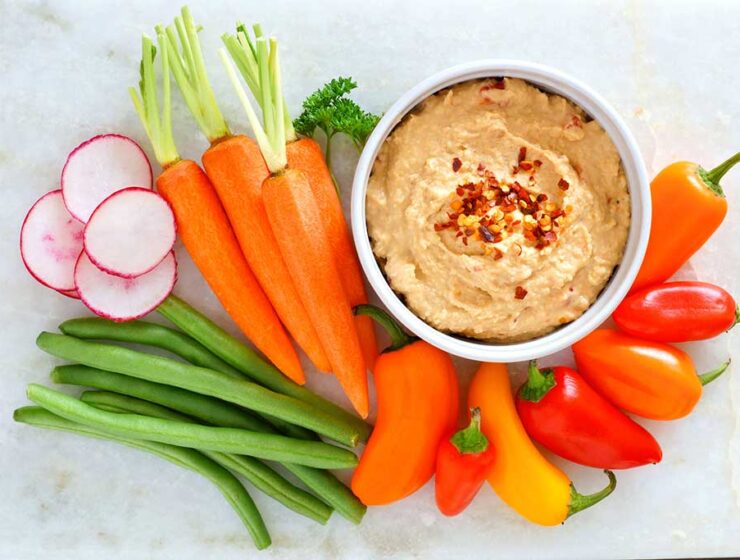 A bowl of hummus topped with red pepper flakes, surrounded by fresh radishes, carrots, green beans, and colorful mini bell peppers.