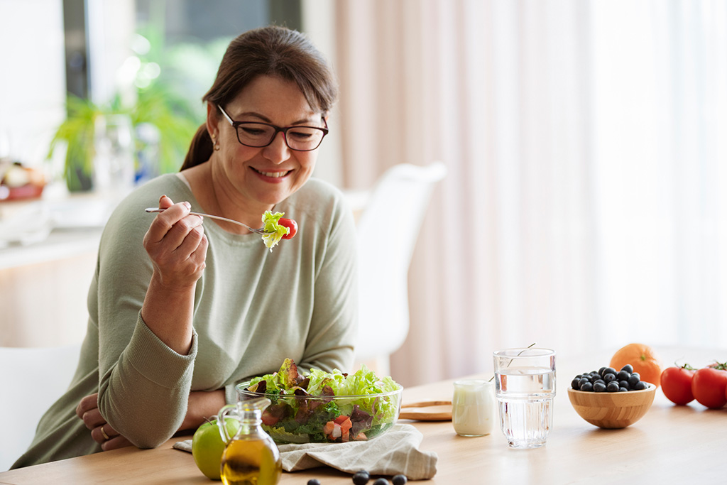 Smiling woman enjoying a fresh salad at a dining table, surrounded by healthy ingredients like fruits, vegetables, and water.