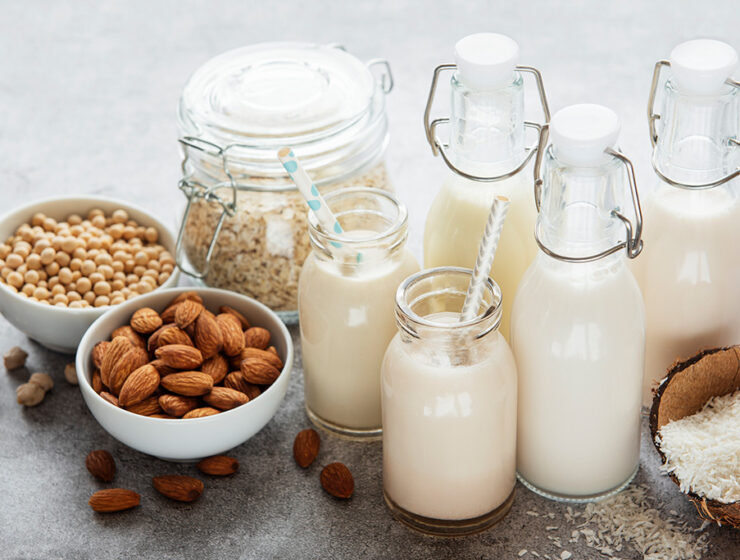 Assorted plant-based milk alternatives in bottles surrounded by almonds, soybeans, oats, and coconut on a gray countertop.