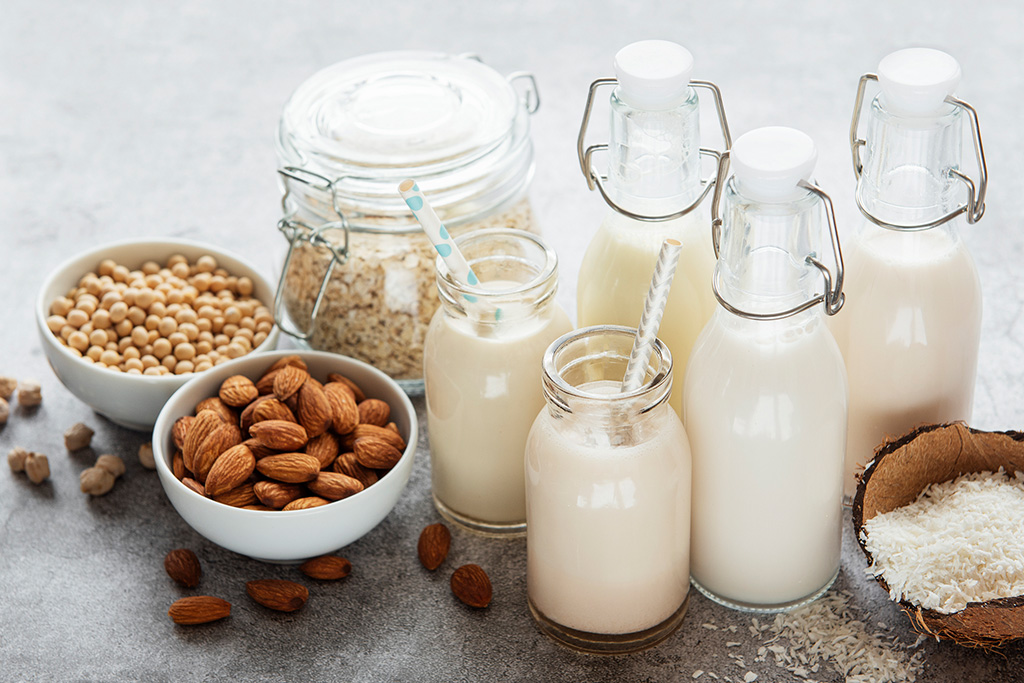 Assorted plant-based milk alternatives in bottles surrounded by almonds, soybeans, oats, and coconut on a gray countertop.
