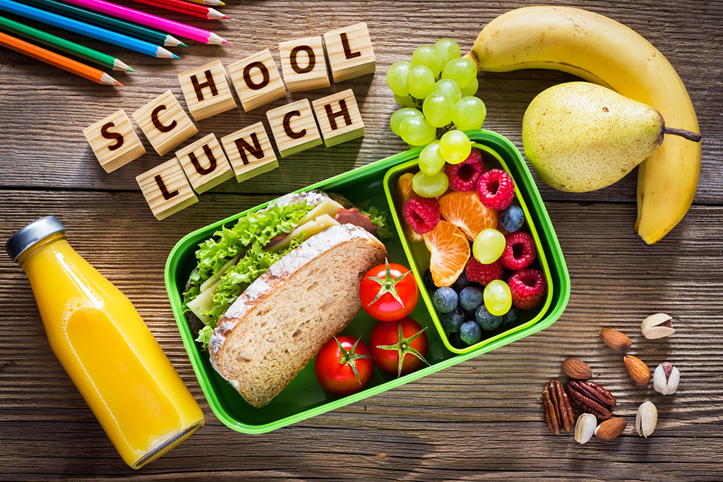 Healthy school lunch with sandwich, fruits, nuts, juice in green lunchbox, and 'School Lunch' in wooden blocks.