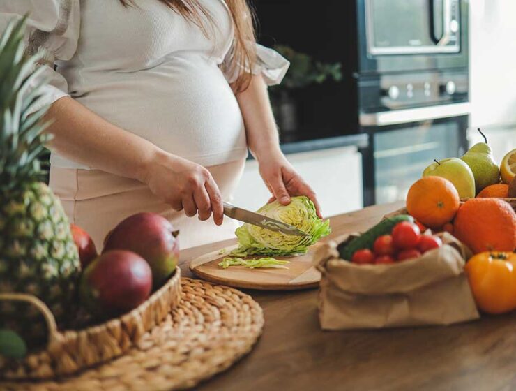 Pregnant woman slicing lettuce in a modern kitchen, surrounded by fresh fruits and vegetables on a wooden table.