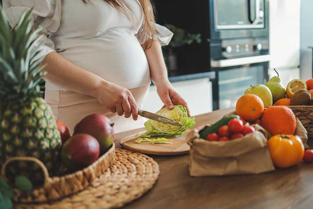 Pregnant woman slicing lettuce in a modern kitchen, surrounded by fresh fruits and vegetables on a wooden table.
