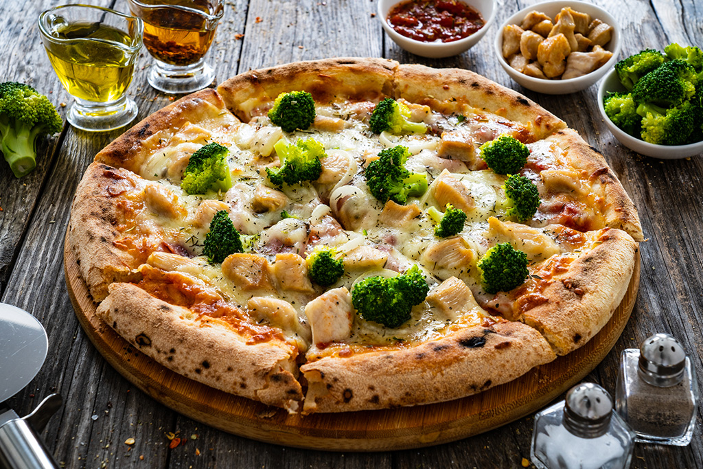 Delicious pizza topped with broccoli and chicken on a wooden table, accompanied by olive oil, chili flakes, and side dishes.